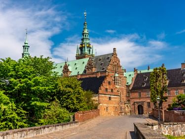 Schloss Frederiksborg in Hillerød mit roten Backsteingebäuden, grünen Kupfertürmen und einer Steinbrücke zum Eingang unter blauem Himmel.
