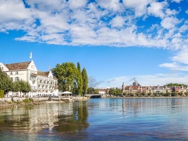 View across Lake Constance showing a white historic building on the left shore and the old town of Konstanz on the right under a blue sky.