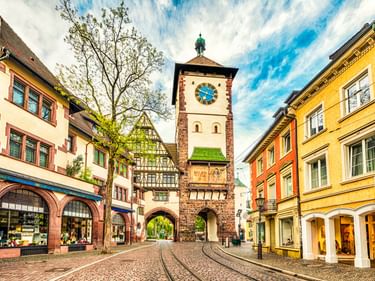 Historisches Schwabentor mit Uhr in Freiburg, umgeben von bunten Fachwerkhäusern an einer Kopfsteinpflasterstraße unter blauem Himmel.