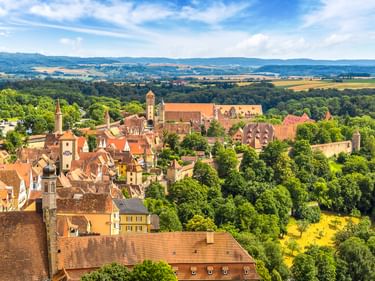 Aerial view of Rothenburg with medieval buildings, red-tiled roofs, church towers, and green forests. Rolling countryside visible in background.