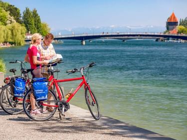 Zwei Radfahrer mit Rädern lesen eine Karte am Seerheinufer. Eine Brücke und ein Turm sind über dem Wasser sichtbar, Berge im Hintergrund.