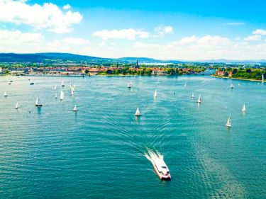 Aerial view of Lake Constance with numerous sailboats and a motorboat creating a wake. The city of Konstanz is visible in the background.