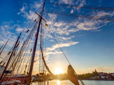 Segelschiffmasten im Eckernförder Hafen bei Sonnenuntergang mit blauem Himmel, goldener Sonne auf dem Wasser und bunten Häusern am Ufer.