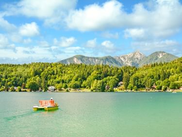 Gelb-grünes Tretboot auf dem türkisfarbenen Klopeiner See mit bewaldeten Ufern und Bergkette unter blauem Himmel mit weißen Wolken in Österreich.