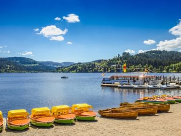 Bunte Tretboote und Ruderboote am Sandstrand des Titisees. Fahrgastschiff am Steg, bewaldete Hügel im Hintergrund unter blauem Himmel.