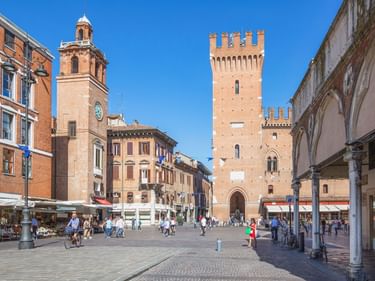 Sonniger Platz in Ferrara mit zinnenbekröntem mittelalterlichem Turm, Glockenturm und historischen Gebäuden. Fußgänger auf dem Kopfsteinpflaster.