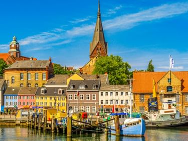 Bunte historische Gebäude säumen den Flensburger Hafen mit Booten an Holzstegen. Der hohe Turm der Marienkirche ragt über die Stadt.