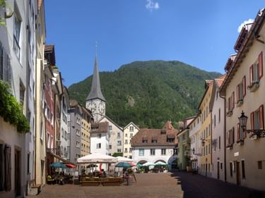 Fußgängerplatz in Churs Altstadt mit bunten Gebäuden, Kirchturm, Straßencafés und grüner Bergkulisse unter blauem Himmel.