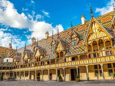 Innenhof des Hôtel-Dieu Museums in Beaune mit buntem glasierten Ziegeldach in geometrischen Mustern, Fachwerk-Giebeln und gotischen Türmen unter blauem Himmel.