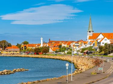 Küstenansicht von Rønne auf Bornholm mit weißem Leuchtturm, Kirche mit orangefarbenem Dach und Turm, bunten Häusern und felsiger Küste am blauen Wasser.