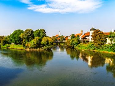 Blick auf Donauwörth mit der Donau im Vordergrund, historischen Gebäuden mit roten Dächern, Kirchtürmen und grünen Bäumen unter blauem Himmel.