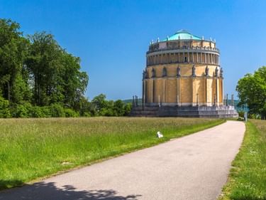 Befreiungshalle monument in Kelheim, a circular neoclassical building with columns, viewed from a paved path through green meadows under blue sky.