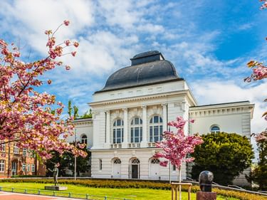 Weißes neoklassizistisches Theatergebäude in Rendsburg mit Kuppeldach, eingerahmt von rosa Kirschblüten und grüner Rasenfläche unter blauem Himmel.