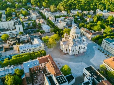 Luftaufnahme des Stadtzentrums von Kaunas mit einer weißen Kuppelkirche, umgeben von einem runden Platz, Wohngebäuden und grünen Bäumen.