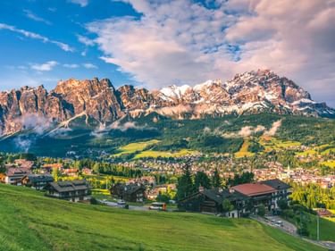 Panoramablick auf Cortina d'Ampezzo im Tal mit dramatischen Dolomitengipfeln dahinter unter blauem Himmel mit Wolken.