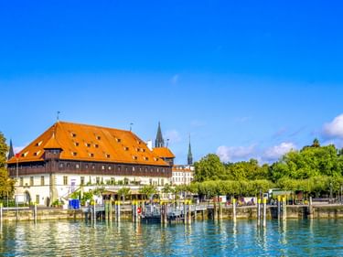 Historisches Konzilgebäude mit orangefarbenem Ziegeldach am Konstanzer Hafen am Bodensee. Holzstege im Vordergrund, Kirchtürme dahinter.