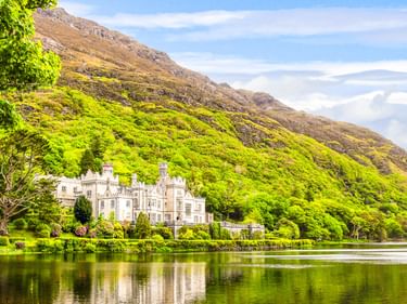 Kylemore Abbey, ein weißes schlossähnliches Gebäude, eingebettet in üppig grüne Bäume neben einem ruhigen See mit felsigem Berg dahinter.