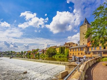 Uferpromenade in Calbe an der Saale mit gelben historischen Gebäuden, Steinturm und Wehr im Fluss unter blauem Himmel mit weißen Wolken.