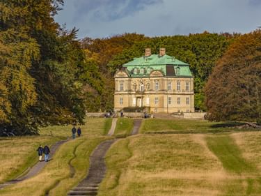 Jagdschloss Eremitageslottet mit gelber Fassade und grünem Kupferdach, umgeben von Herbstbäumen. Geschwungene Wege führen durch Wiesen zum Schloss.