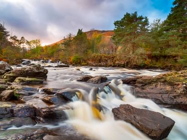 Dochart-Wasserfälle in Killin mit rauschenden Wasser über Felsen. Bäume säumen beide Ufer, Hügel erheben sich im Hintergrund unter bewölktem Himmel.