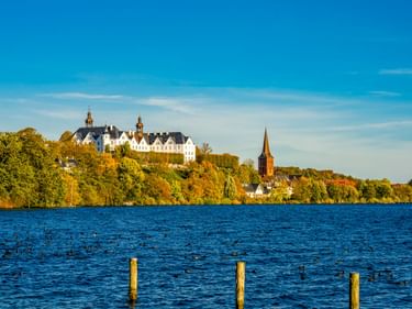 Plöner Schloss, ein weißes Barockschloss, thront auf einem Hügel über einem blauen See im Herbst. Ein Kirchturm und bunte Bäume sind am Ufer sichtbar.