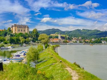Blick auf Grein mit Schloss Greinburg an der Donau. Grüne Uferböschung mit Booten, Ortschaft mit Kirchturm und bewaldete Hügel unter blauem Himmel.
