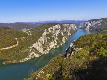 Panoramablick auf die Donau, die sich durch steile Kalksteinfelsen am Eisernen Tor windet. Bewaldete Hügel und eine kurvige Straße sind sichtbar.