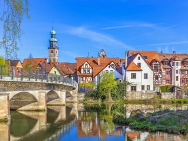 Steinbrücke über die Pegnitz mit bunten Fachwerkhäusern und Kirchturm in Lauf an der Pegnitz, Franken, unter blauem Himmel.