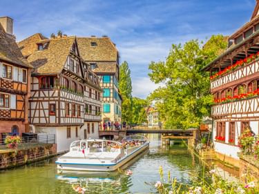Touristenboot auf Kanal in Petite France, Straßburg, mit traditionellen Fachwerkhäusern und Blumenkästen entlang des Wassers unter blauem Himmel.