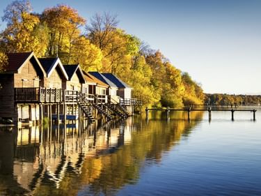 Reihe von Holzbootshäusern auf Stelzen am Ammersee mit Herbstbäumen und Holzsteg. Klarer blauer Himmel und ruhiges Wasser mit Spiegelungen.
