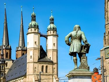 Bronze statue in front of historic church towers in Lutherstadt Wittenberg under clear blue sky. Twin spires and domed towers visible.