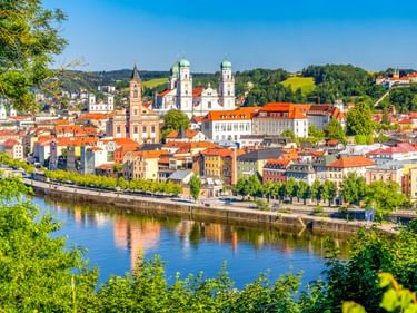 Panoramablick auf Passau mit bunten Gebäuden, Kirchtürmen mit grünen Kuppeln und einem Fluss, der den blauen Himmel spiegelt. Grüne Hügel im Hintergrund.