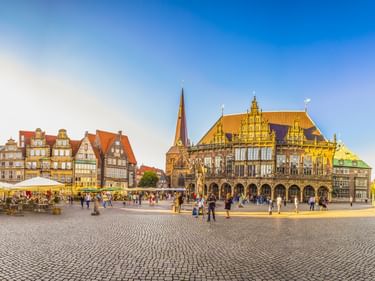 Bremen Town Hall Square with ornate Renaissance town hall, church spire, and colorful historic buildings. Cobblestone plaza with visitors and cafes.