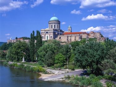 Basilika von Esztergom mit türkisfarbener Kuppel auf einem Hügel mit Blick auf die Donau. Historische Festungsmauern und üppige grüne Bäume umgeben das Flussufer.