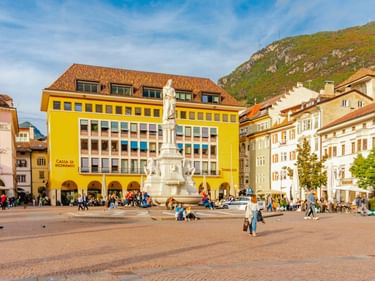 Waltherplatz in Bozen mit weißer Statue in der Mitte, gelbem Gebäude mit Arkaden und Bergkulisse. Menschen flanieren über den Platz.