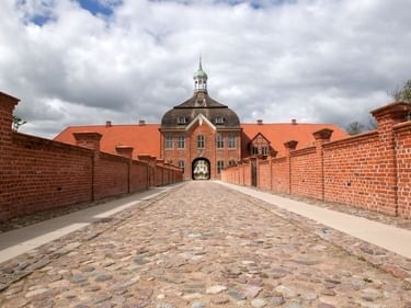 Kopfsteinpflasterweg zu einem barocken Torhaus mit roten Backsteinmauern, orangefarbenem Ziegeldach und grüner Kupferkuppel unter bewölktem Himmel.