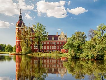 Red baroque Schloss Muskau in Bad Muskau with tower and white gables, surrounded by green trees, reflected in calm lake water under blue sky.