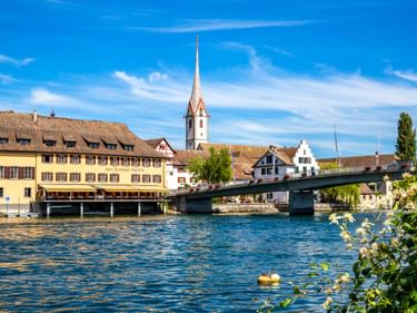 View of Stein am Rhein waterfront with historic buildings, a church with tall white spire, and a bridge crossing the Rhine River under blue sky.