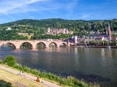 Old stone bridge crossing the Neckar River in Heidelberg with castle ruins on hillside, church tower, and historic buildings along the riverbank.
