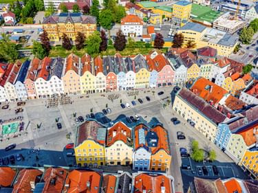 Luftaufnahme des historischen Stadtplatzes von Schärding mit bunten Barockgebäuden in Gelb, Orange und Blau um einen großen Platz mit parkenden Autos.