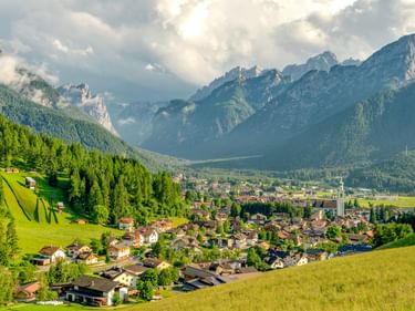 Toblach liegt in einem grünen Tal, umgeben von bewaldeten Hügeln und dramatischen Dolomitengipfeln unter bewölktem Himmel.