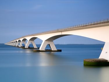 Lange weiße Zeelandbrücke mit markanten Bogenpfeilern, die sich über ruhiges blaues Wasser unter klarem Himmel in den Niederlanden erstreckt.