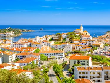 Luftaufnahme von Cadaqués an der Costa Brava mit weißen Häusern, orangefarbenen Dächern und einer Kirche mit Blick auf das blaue Mittelmeer.
