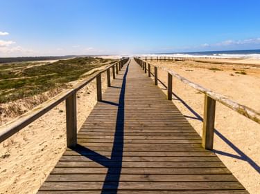 Langer Holzsteg mit Geländer führt über Sanddünen am Strand von Furadouro. Meereswellen in der Ferne unter blauem Himmel sichtbar.