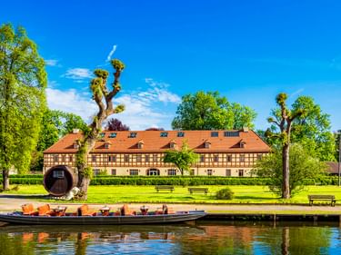 Traditionelle Boote am Großen Spreehafen in Lübbenau. Fachwerkgebäude mit orangem Dach hinter grüner Wiese und hohen Bäumen unter blauem Himmel.
