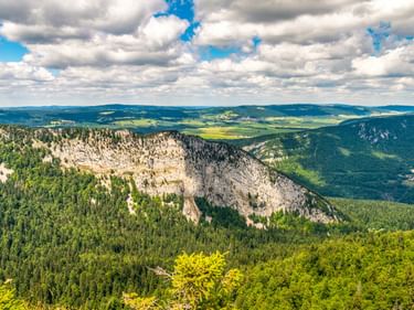 Luftaufnahme des Val de Travers mit Kalksteinfelsen, dichten Wäldern und grünem Tal unter blauem Himmel mit weißen Wolken.