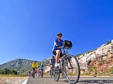 Radfahrer in blauem Trikot auf asphaltierter Straße in Dalmatien mit Felsklippen und Hügeln. Radfahrergruppe im Hintergrund unter blauem Himmel.