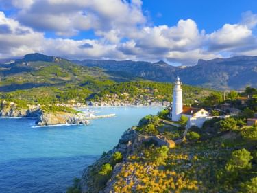 Weißer Leuchtturm auf felsiger Klippe mit Blick auf die Bucht von Port de Sóller mit türkisfarbenem Wasser, Küstenort und Tramuntana-Bergen.