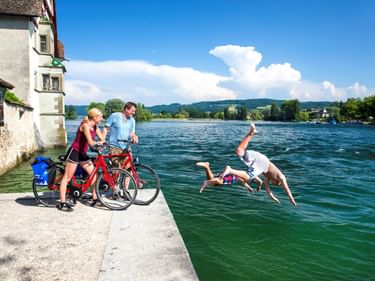 Zwei Radfahrer mit roten Rädern auf einer Uferpromenade in Stein am Rhein beobachten zwei Personen beim Sprung in den Rhein. Historisches Gebäude sichtbar.