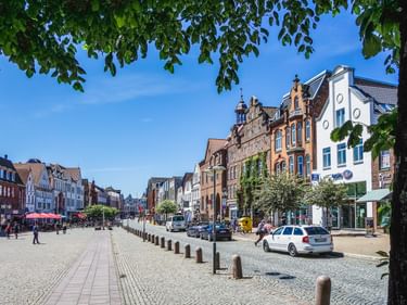 Kopfsteinpflaster-Marktplatz in Husum Altstadt mit bunten historischen Gebäuden, parkenden Autos und Bäumen unter blauem Himmel.